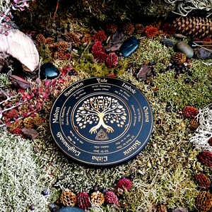 May include: A black wooden wheel with gold lettering and a gold tree of life design. The wheel is labeled with the eight sabbats of the Wheel of the Year: Samhain, Mabon, Litha, Lughnasadh, Beltane, Ostara, Imbolc, and Yule.