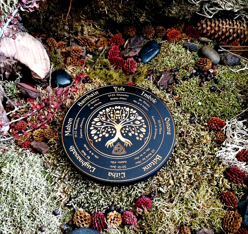 May include: A black wooden wheel with gold lettering and a gold tree of life design. The wheel is labeled with the eight sabbats of the Wheel of the Year: Samhain, Mabon, Litha, Lughnasadh, Beltane, Ostara, Imbolc, and Yule.