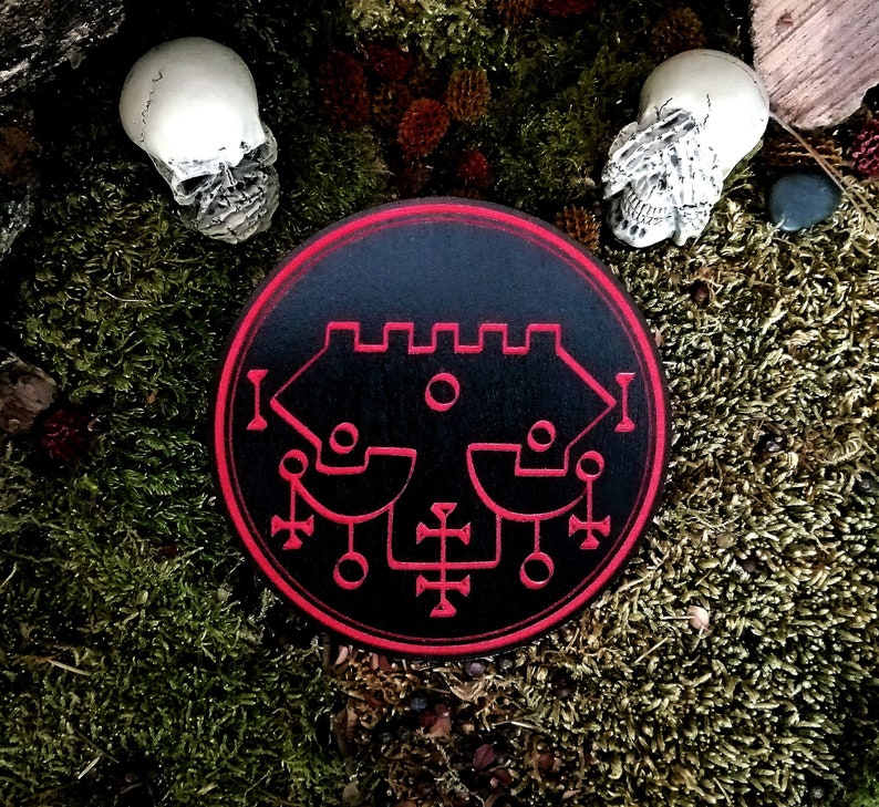May include: A black and red circular sigil with a complex geometric design. The sigil is surrounded by a red border and is set against a mossy green background.