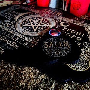 May include: A black wooden Ouija board with gold lettering and a pentagram design. The board includes numbers 1-9, letters A-Z, and the words "Good Bye" and "Ouija". The board is on a brown surface with a red and black candle in the background.