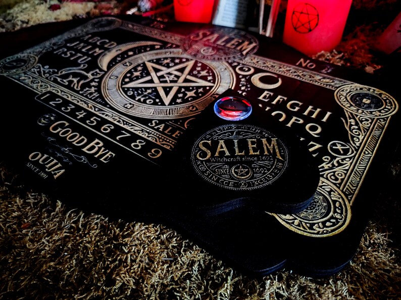 May include: A black wooden Ouija board with gold lettering and a pentagram design. The board includes numbers 1-9, letters A-Z, and the words "Good Bye" and "Ouija". The board is on a brown surface with a red and black candle in the background.