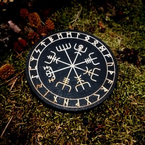 May include: A black circular wooden compass with a white compass rose and runes around the edge. The compass is on a bed of green moss.