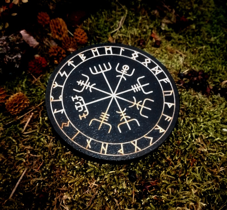 May include: A black circular wooden compass with a white compass rose and runes around the edge. The compass is on a bed of green moss.