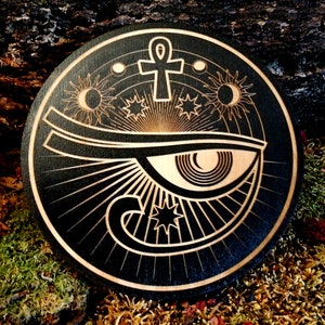 May include: A black wooden round plaque with a laser-etched design of the Eye of Horus, an ankh symbol, and celestial bodies like the sun, moon, and stars.