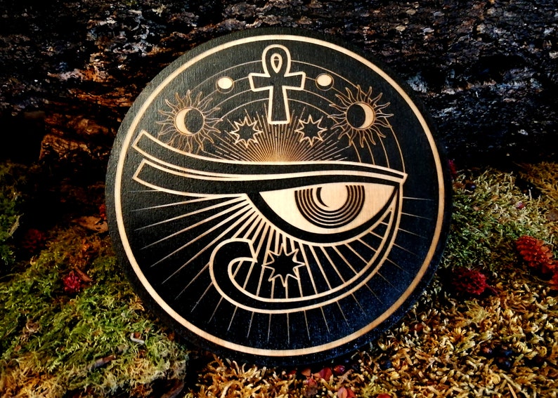 May include: A black wooden round plaque with a laser-etched design of the Eye of Horus, an ankh symbol, and celestial bodies like the sun, moon, and stars.