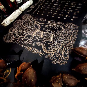 May include: A black and gold witch's alphabet chart with a tree and snake design. The chart includes the letters of the alphabet and symbols.