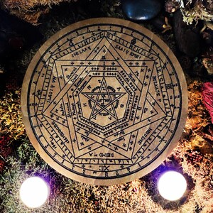 May include: A wooden circle with a black and white pentagram design. The circle is surrounded by a ring of text in a circular pattern. The text includes the words "XE", "DEIMO A", "126 MEGCBX", "ILAGIV VN", "SAATY EME", "BIZKASE", "MIGAA", "IHI LAAY", and "ZIIRKS".