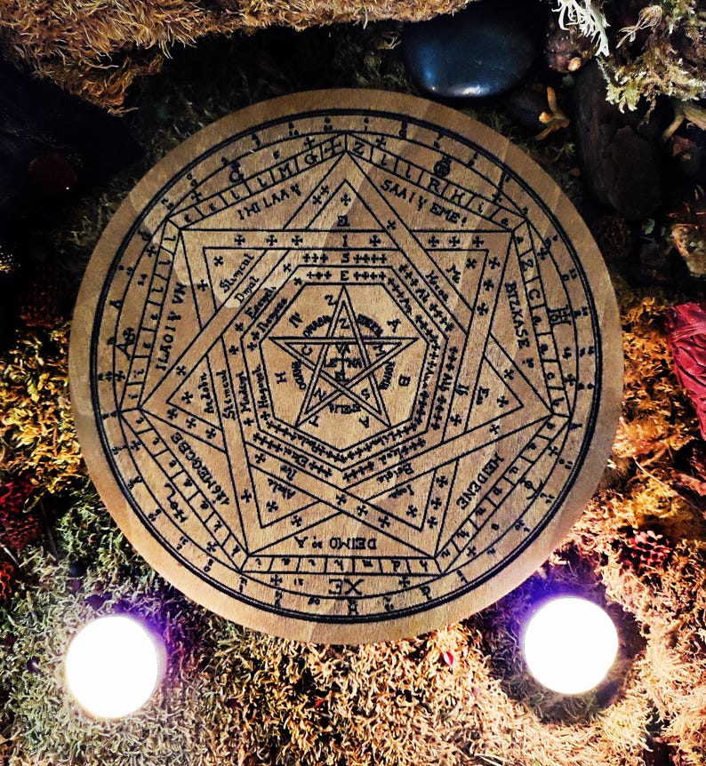 May include: A wooden circle with a black and white pentagram design. The circle is surrounded by a ring of text in a circular pattern. The text includes the words "XE", "DEIMO A", "126 MEGCBX", "ILAGIV VN", "SAATY EME", "BIZKASE", "MIGAA", "IHI LAAY", and "ZIIRKS".