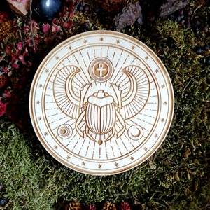 Peut inclure: Un dessous de verre rond en bois avec un motif gravé au laser d'un scarabée aux ailes déployées. Le scarabée est entouré d'un cercle d'étoiles et d'un soleil, d'une lune et d'un symbole Ankh.