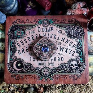 May include: A wooden Ouija board with an ornate design featuring a crescent moon, a sun, and a skull. The board has a planchette with a blue eye in the center. The letters of the alphabet, numbers 1-9 and 0, and the words "YES", "NO", and "GOOD BYE" are printed on the board.