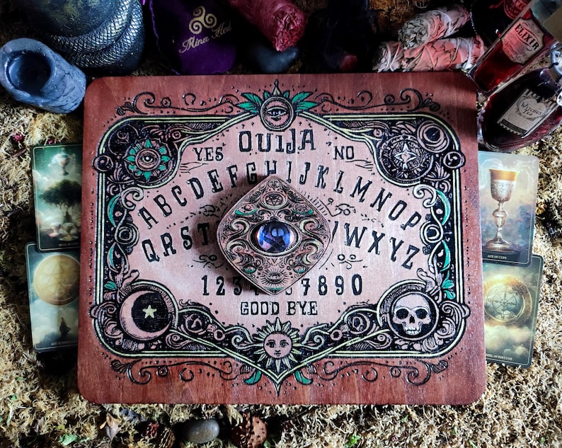 May include: A wooden Ouija board with an ornate design featuring a crescent moon, a sun, and a skull. The board has a planchette with a blue eye in the center. The letters of the alphabet, numbers 1-9 and 0, and the words "YES", "NO", and "GOOD BYE" are printed on the board.