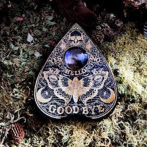 May include: A black wooden Ouija board with a gold-colored design. The board features a moth, a crescent moon, and the words "YES", "NO", "HELLO", and "GOODBYE". A purple iridescent glass orb sits in the center of the board.