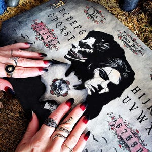 May include: A grey Ouija board with black and red details, depicting a skull and a woman's face. The board displays the alphabet, numbers, and the words "Yes" and "Good Bye." The image also shows hands with black and red ombre nails and various rings.