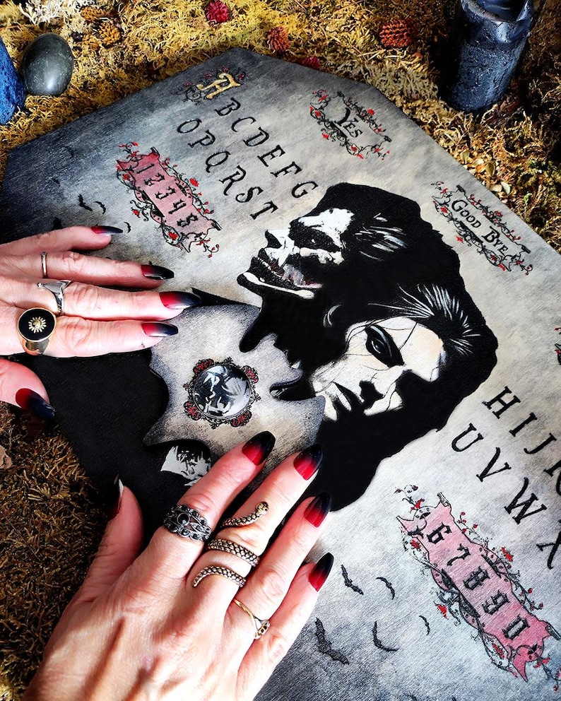 May include: A grey Ouija board with black and red details, depicting a skull and a woman's face. The board displays the alphabet, numbers, and the words "Yes" and "Good Bye." The image also shows hands with black and red ombre nails and various rings.
