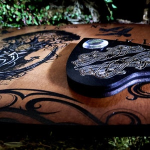 May include: A wooden Ouija board with a black and gold design. The board features a heart-shaped planchette with a clear glass lens. The board is surrounded by a dark green mossy background.