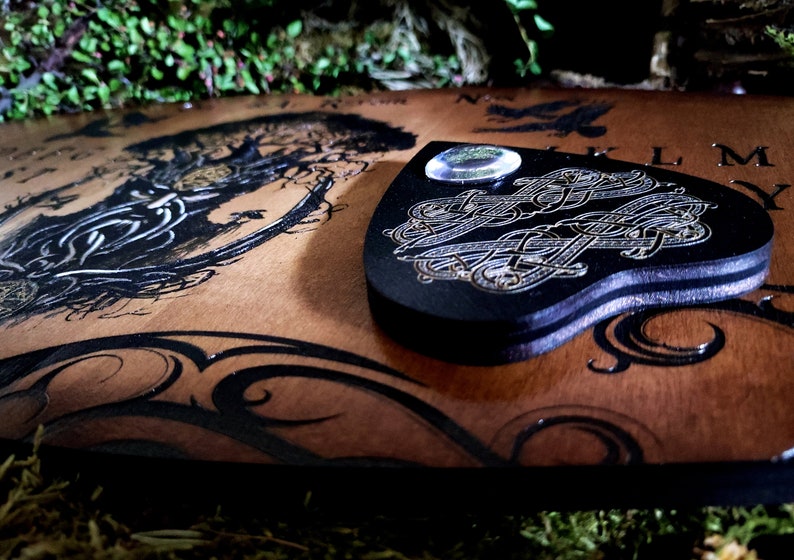 May include: A wooden Ouija board with a black and gold design. The board features a heart-shaped planchette with a clear glass lens. The board is surrounded by a dark green mossy background.