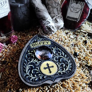 May include: A black and gold planchette with the name "BUFFY" on top. The planchette has a large crystal and a gold cross design. The planchette is surrounded by other mystical items, including bottles and sage bundles.
