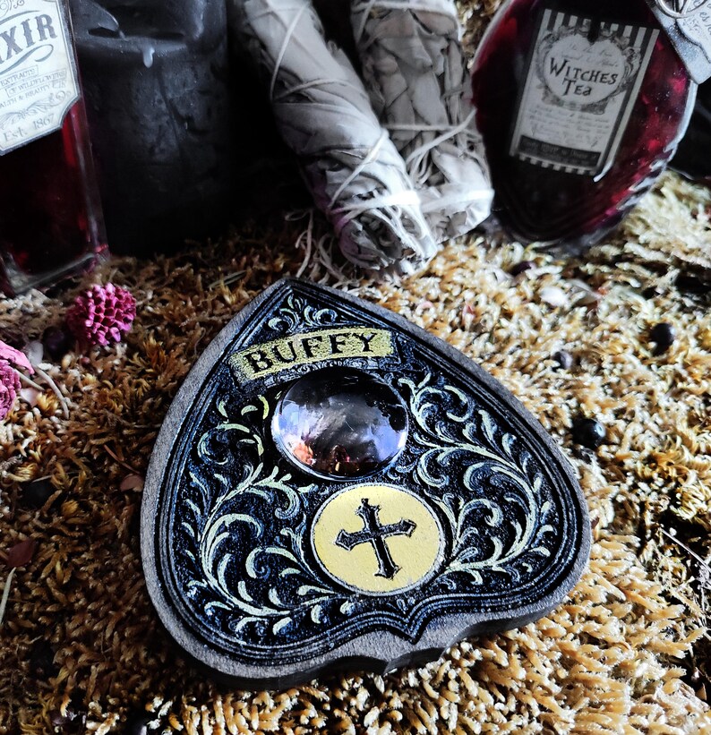 May include: A black and gold planchette with the name "BUFFY" on top. The planchette has a large crystal and a gold cross design. The planchette is surrounded by other mystical items, including bottles and sage bundles.