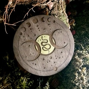 May include: A round wooden coaster with a triple moon and snake design. The coaster is dark brown with a gold snake and moon design. The coaster is surrounded by a ring of small stars.