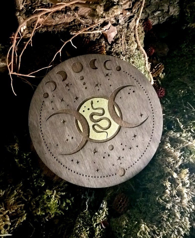 May include: A round wooden coaster with a triple moon and snake design. The coaster is dark brown with a gold snake and moon design. The coaster is surrounded by a ring of small stars.