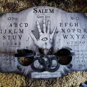 K&ouml;nnte beinhalten: Ein Ouija-Brett aus Holz mit den Worten "SALEM" und "GOOD BYE" darauf gedruckt. Das Brett hat ein schwarz-wei&szlig;es Alphabet, Zahlen und eine Hand mit einem Auge in der Mitte. Das Brett ist von Moos umgeben.