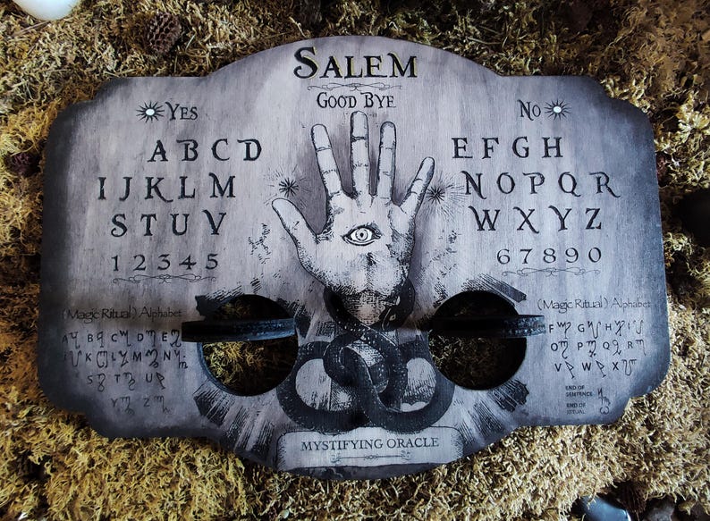 K&ouml;nnte beinhalten: Ein Ouija-Brett aus Holz mit den Worten "SALEM" und "GOOD BYE" darauf gedruckt. Das Brett hat ein schwarz-wei&szlig;es Alphabet, Zahlen und eine Hand mit einem Auge in der Mitte. Das Brett ist von Moos umgeben.