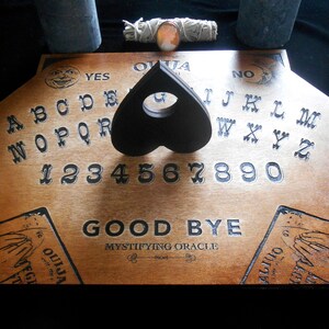 May include: Wooden Ouija board with a black heart-shaped planchette, the letters A through Z, numbers 1 through 9 and 0, and the words "YES", "NO", and "GOOD BYE".