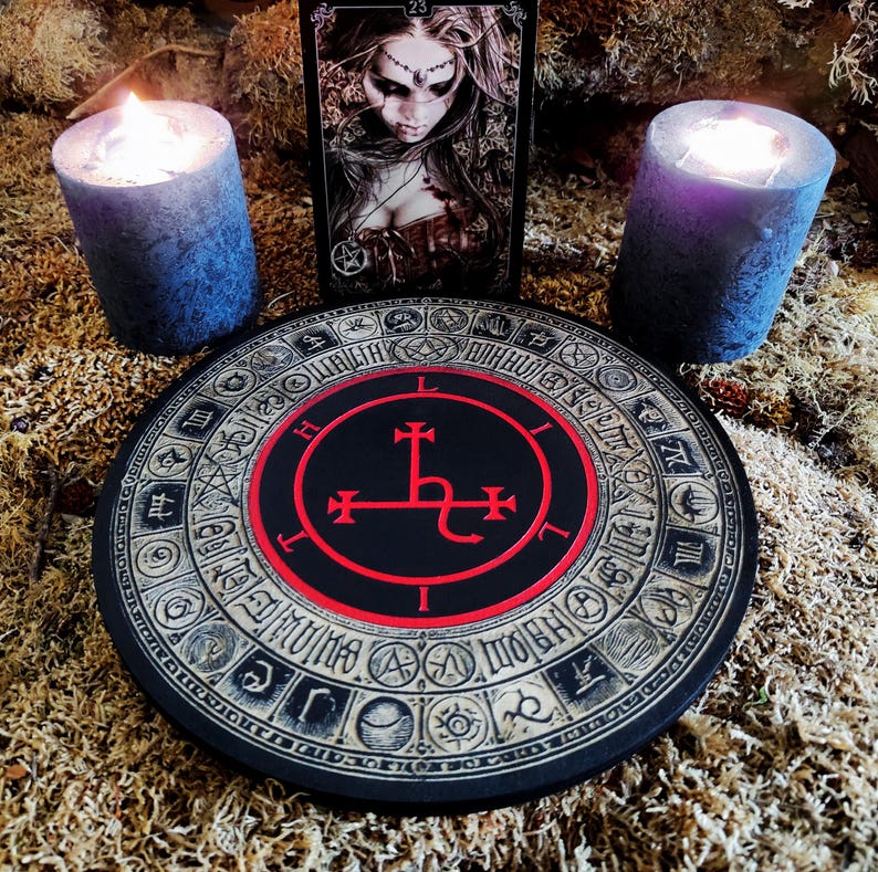 May include: A black and red circular wooden board with a sigil in the center. The sigil is a black cross with a snake wrapped around the base. The board is surrounded by a circle of symbols and numbers. Two black candles are on either side of the board.
