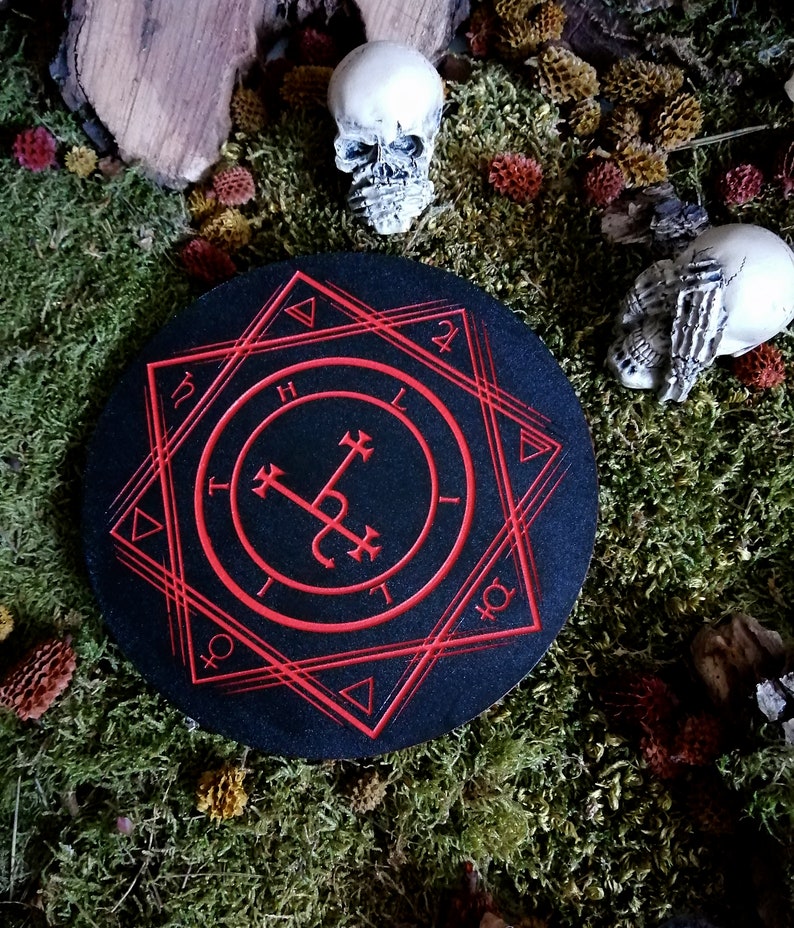 Op de afbeelding: Een zwarte ronde onderzetter met een rood zegel in het midden. Het zegel wordt omringd door een geometrisch patroon van driehoeken en cirkels. De onderzetter ligt op een bed van groen mos.