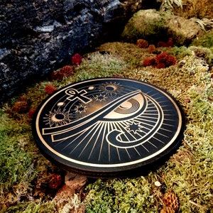 May include: A black wooden round plaque with a gold design of an eye, an ankh, and stars. The design is surrounded by rays of light.
