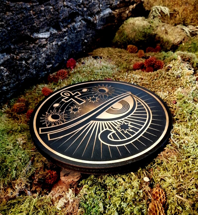 May include: A black wooden round plaque with a gold design of an eye, an ankh, and stars. The design is surrounded by rays of light.