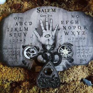 K&ouml;nnte beinhalten: Ein Ouija-Brett aus Holz mit einem schwarz-wei&szlig;en Design. Das Brett enth&auml;lt die Buchstaben des Alphabets, die Zahlen 1-9 und 0 sowie die W&ouml;rter "YES", "NO" und "GOOD BYE". Das Brett zeigt au&szlig;erdem eine Hand mit einem Auge in der Handfl&auml;che, einen Halbmond und eine Sonne. Ein schwarzes und silbernes Planchette befindet sich im Vordergrund.