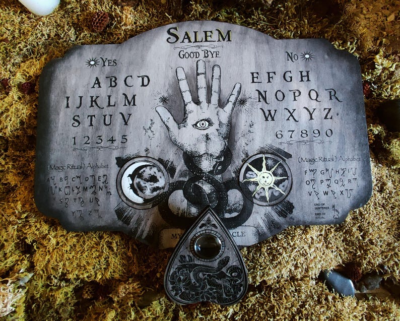 K&ouml;nnte beinhalten: Ein Ouija-Brett aus Holz mit einem schwarz-wei&szlig;en Design. Das Brett enth&auml;lt die Buchstaben des Alphabets, die Zahlen 1-9 und 0 sowie die W&ouml;rter "YES", "NO" und "GOOD BYE". Das Brett zeigt au&szlig;erdem eine Hand mit einem Auge in der Handfl&auml;che, einen Halbmond und eine Sonne. Ein schwarzes und silbernes Planchette befindet sich im Vordergrund.