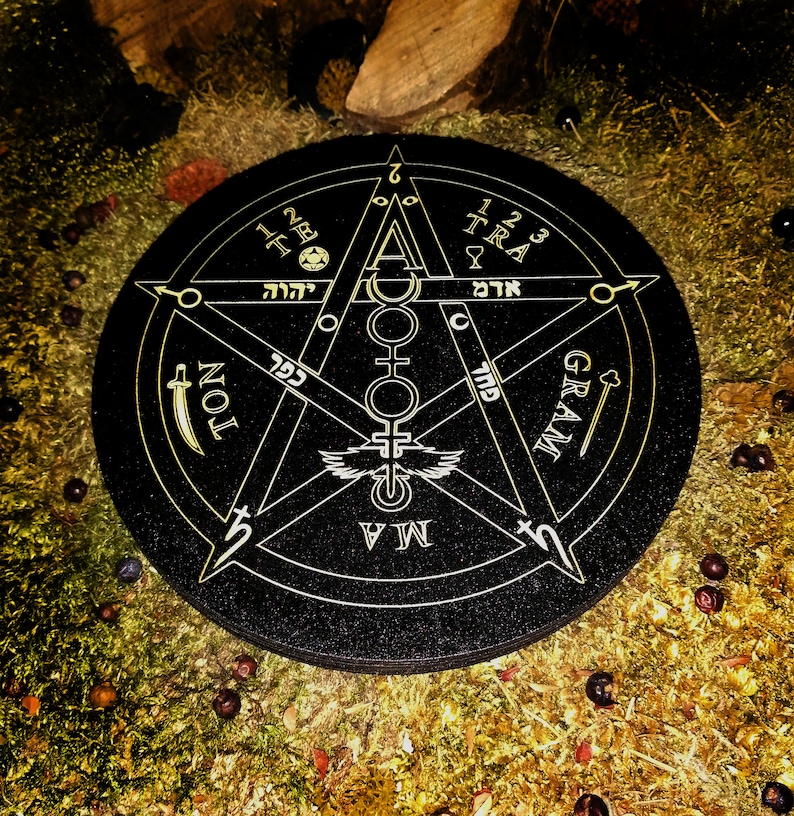 May include: A black circular pentagram with gold lettering and symbols. The pentagram has five points with the words "GRAM", "TRA", "TON", "MAM", and "TEL" inscribed on each point. The centre of the pentagram has a circle with a winged symbol and a series of symbols above and below it. The circle also has the Hebrew words "אד" and "יהוה" inscribed on it.