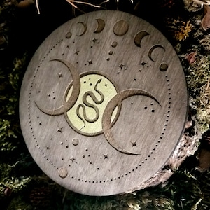 May include: A round wooden coaster with a triple moon design and a snake in the center. The coaster is decorated with stars and moons.