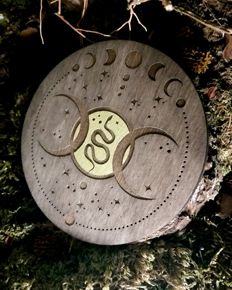 May include: A round wooden coaster with a triple moon design and a snake in the center. The coaster is decorated with stars and moons.