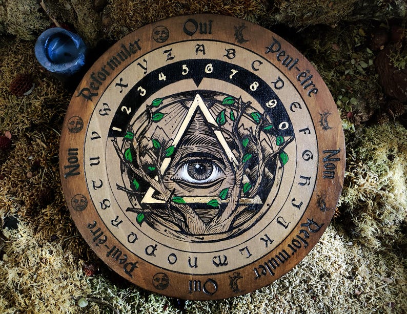 May include: A wooden Ouija board with a gold and black all-seeing eye design. The board has letters, numbers, and the words "Oui", "Peut-&ecirc;tre", "Non", and "Reformuler" around the edge.