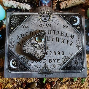 May include: A gray Ouija board with the text "OUIJA SINCE 1891" and a planchette. The board features the alphabet, numbers, and the words "YES" and "NO". The planchette has an eye design. The board is surrounded by tarot cards, candles, and dried moss.