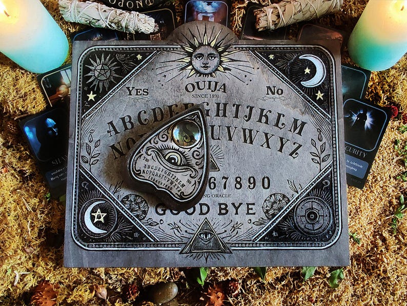 May include: A gray Ouija board with the text "OUIJA SINCE 1891" and a planchette. The board features the alphabet, numbers, and the words "YES" and "NO". The planchette has an eye design. The board is surrounded by tarot cards, candles, and dried moss.