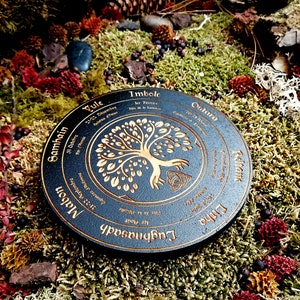 May include: A black wooden wheel with gold lettering and a tree of life design. The wheel is divided into eight sections, each representing a different time of year in the Celtic calendar. The text on the wheel includes the names of the eight festivals: Yule, Samhain, Mabon, Lughnasadh, Litha, Ostara, Imbolc, and Beltane.