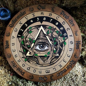 May include: A wooden Ouija board with a gold and black all-seeing eye design. The board has letters, numbers, and the words "Oui", "Peut-&ecirc;tre", "Non", and "Reformuler" around the edge.