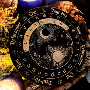 May include: A black circular wooden board with gold lettering and a decorative design. The board features a sun and moon design with a circular scale around the outside. The text on the board reads "Peut-&ecirc;tre Non Reformuler Qui Peut-&ecirc;tre".