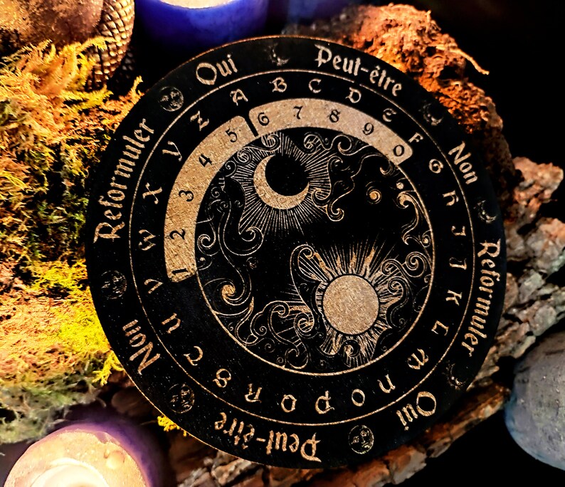 May include: A black circular wooden board with gold lettering and a decorative design. The board features a sun and moon design with a circular scale around the outside. The text on the board reads "Peut-&ecirc;tre Non Reformuler Qui Peut-&ecirc;tre".