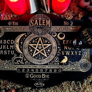 Könnte beinhalten: Ein schwarzes Ouija-Brett aus Holz mit goldenen Buchstaben und einem Pentagramm-Design. Das Brett enthält das Alphabet, die Zahlen 1-9 und 0 sowie die Wörter "Ja", "Nein", "Auf Wiedersehen" und "Ouija seit 1901".