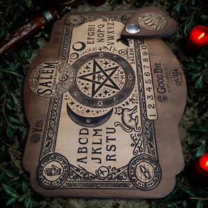May include: Wooden Ouija board with a pentagram, letters, numbers, and the word "Salem" printed on it. A planchette rests on the board. The board is surrounded by lit red candles and greenery.