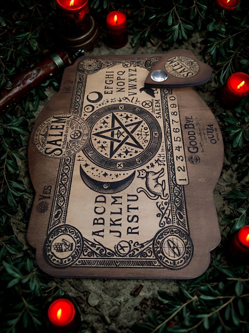 May include: Wooden Ouija board with a pentagram, letters, numbers, and the word "Salem" printed on it. A planchette rests on the board. The board is surrounded by lit red candles and greenery.