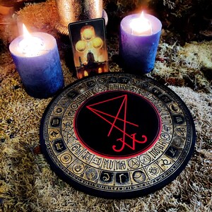Pu&ograve; includere: Un tabellone circolare nero con un bordo dorato e un sigillo rosso al centro. Il tabellone &egrave; circondato da un cerchio di simboli dorati. Due candele blu sono accese su entrambi i lati del tabellone. Una carta dei tarocchi con uno sfondo dorato e tre sfere dorate &egrave; visibile dietro il tabellone.