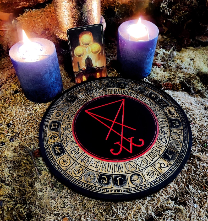 Pu&ograve; includere: Un tabellone circolare nero con un bordo dorato e un sigillo rosso al centro. Il tabellone &egrave; circondato da un cerchio di simboli dorati. Due candele blu sono accese su entrambi i lati del tabellone. Una carta dei tarocchi con uno sfondo dorato e tre sfere dorate &egrave; visibile dietro il tabellone.