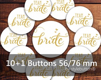 May include: White round buttons with gold text that reads "TEAM BRIDE". The buttons are scattered on a wooden surface. The text "10+1 Buttons 56/76 mm" is also visible.