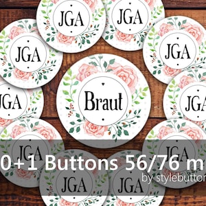 Könnte beinhalten: Elf runde, weiße, floral gemusterte Knöpfe mit dem Text "JGA" in Schwarz. Ein Knopf hat den Text "Braut" in Schwarz. Die Knöpfe haben einen Durchmesser von 56/76 mm.